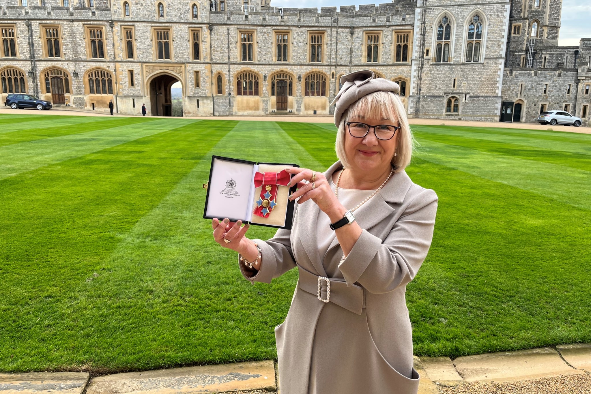 GPA Chair Pat Richie with her CBE award in her hands, Pat is smiling whilst holding the award, standing in front of Windsor Castle and the green grassed lawn. Pat is wearing a grey jacket and hat. 
