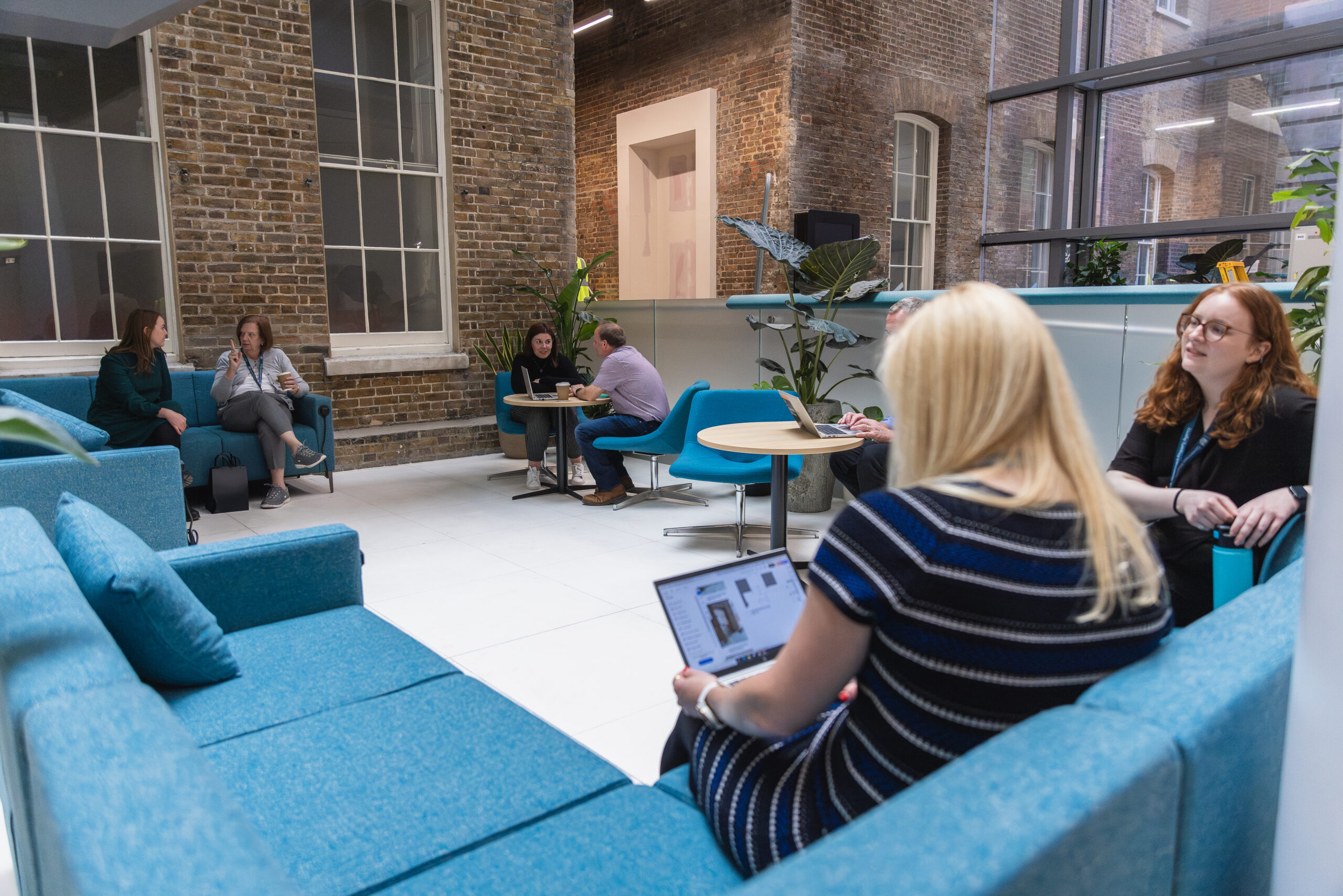 Colleague sitting on a sofa working on laptop. The sofa is bright blue and positioned in a touchdown space.