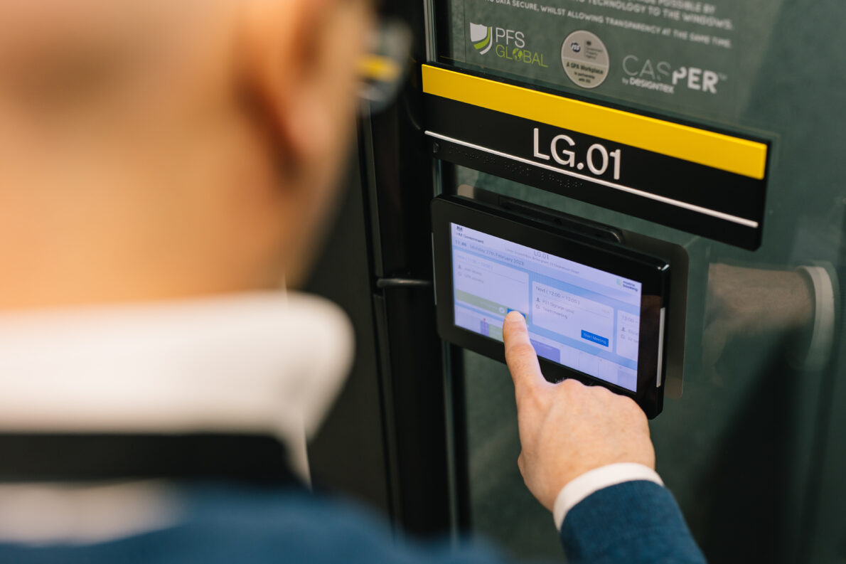 A person holds their finger against a meeting room booking panel, lit up on the wall