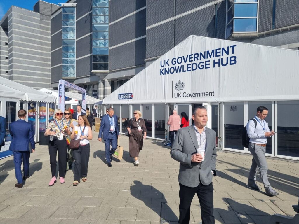 An outdoor shot on a sunny day shows people walking in a paved plaza in front of a large white marquee labeled "UK GOVERNMENT KNOWLEDGE HUB" and "UK Government." A smaller "UKREiiF" logo is also visible on the tent. Several people are wearing lanyards and business-casual attire, some carrying coffee or bags, suggesting a professional conference or event. In the background is a large, modern grey building with glass window columns.