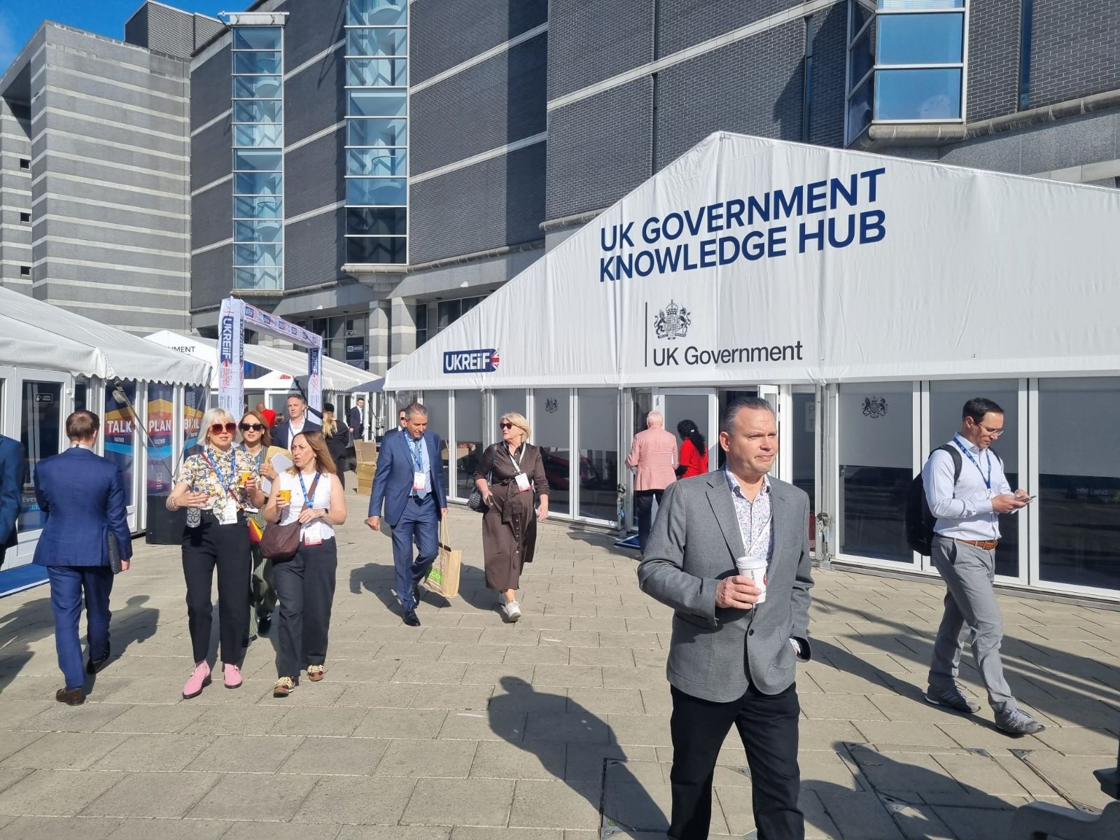An outdoor shot on a sunny day shows people walking in a paved plaza in front of a large white marquee labeled "UK GOVERNMENT KNOWLEDGE HUB" and "UK Government." A smaller "UKREiiF" logo is also visible on the tent. Several people are wearing lanyards and business-casual attire, some carrying coffee or bags, suggesting a professional conference or event. In the background is a large, modern grey building with glass window columns.