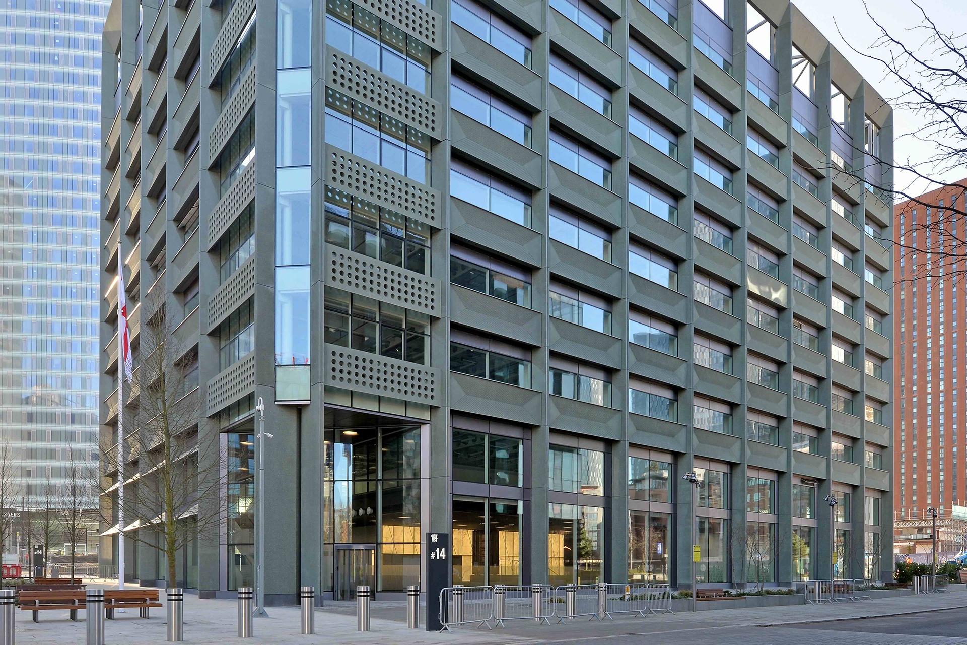 An exterior, close-up view of an eight-story modern office building in Manchester, England, featuring a dark green/grey perforated metal facade and large horizontal rectangular windows. The ground floor has a recessed glass entrance and the building is set back from a paved public space with bollards and benches.