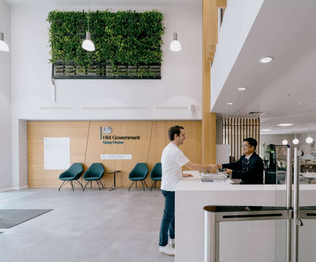 A man hands his GovPass to reception staff at Quay House, Peterborough. In the foreground are security gates with GovPass scanner terminals.